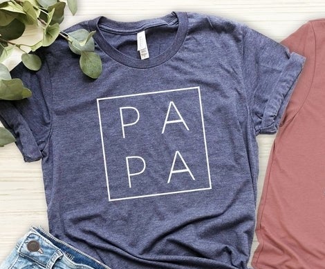 A heather blue t-shirt with a white square and the two sets of the letters PA stacked on top of each other