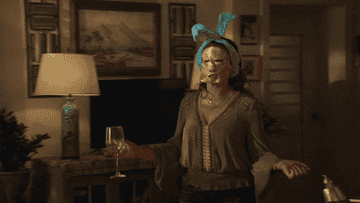 gif of a person dancing through their living room with a face mask on their face their hair pushed back behind their ears with a spa headband and a glass of wine