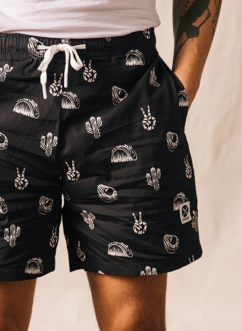 black shorts with illustrations of tacos, avocados, cacti, and skeleton hand peace signs in white