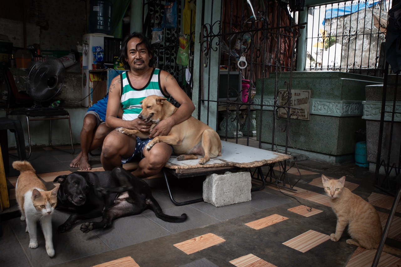 A man with two dogs and two cats sits on a cot in a graveyard in the Philippines 