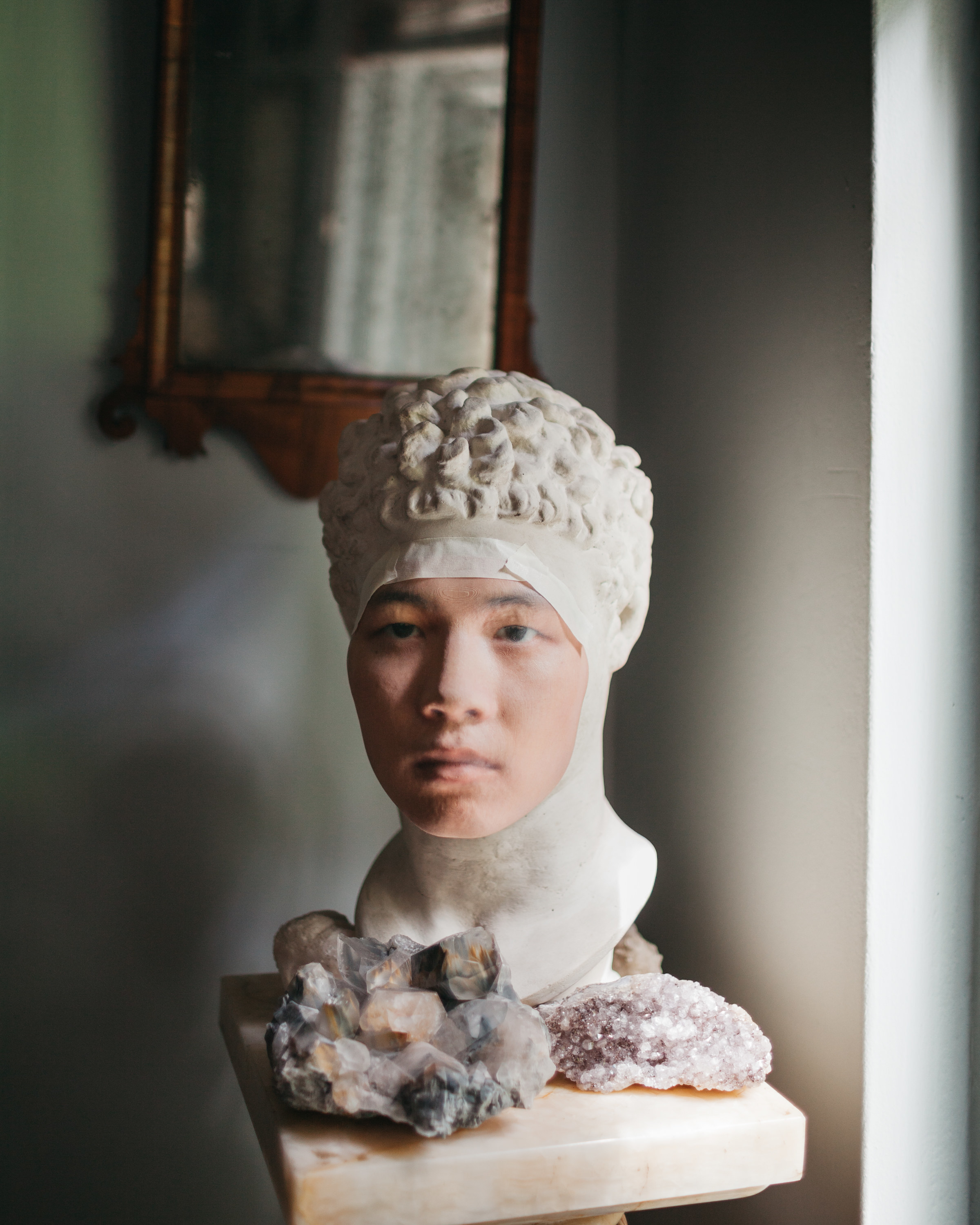 A mans face taped to a bust statue with crystals around it on a pedestal 