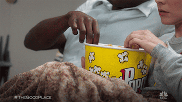 Chidi and Eleanor from &quot;The Good Place&quot; eating popcorn shrimp out of a popcorn bucket and watching TV