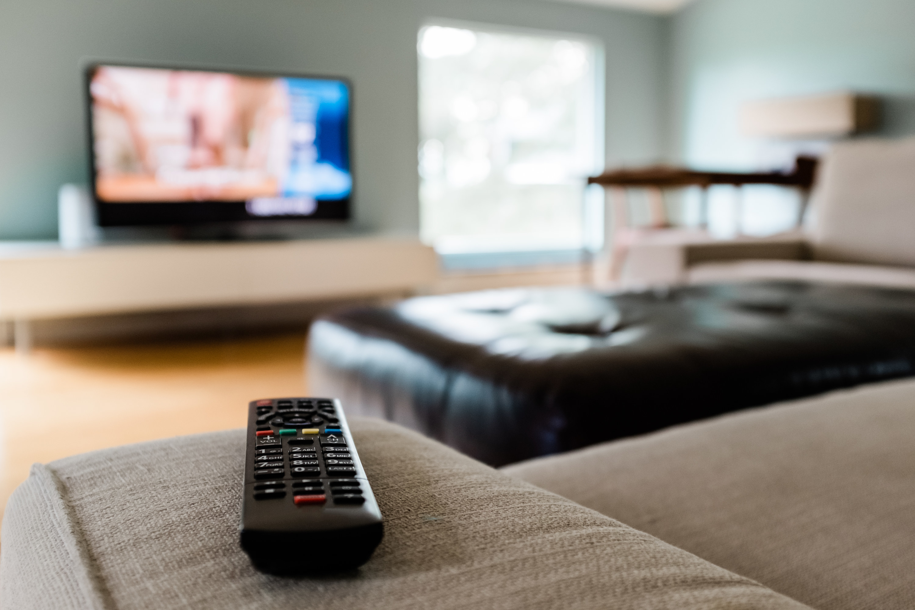 Photo of a living room couch with a tv remote in the forefront and a tv playing in the background