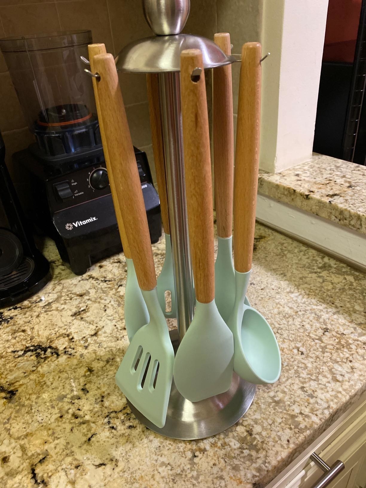 A reviewer photo of the utensils in light green, hanging from their storage stand, which has a circular base, thin body, and hooks at the top that can hold the utensils