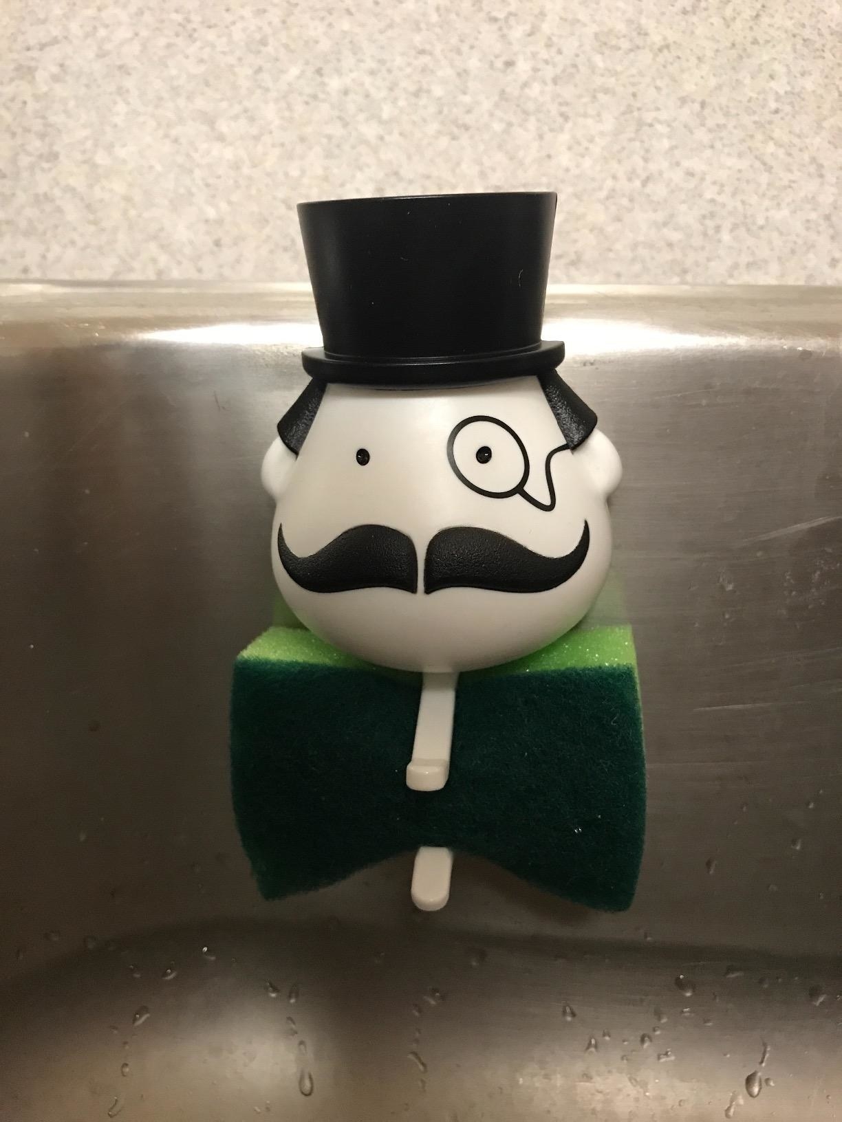 A reviewer photo of the sponge holder, which has a small &quot;head&quot; with a top hat, monocle, and mustache, and which holds the sponge below the head as if it were a bow tie