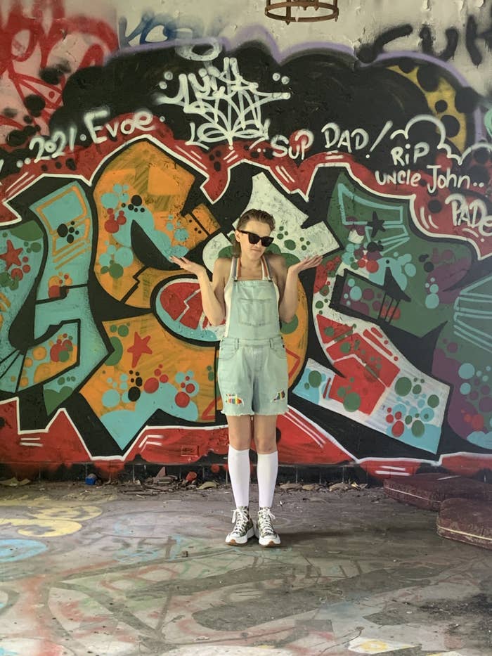 A trans person shrugs before a background of graffiti