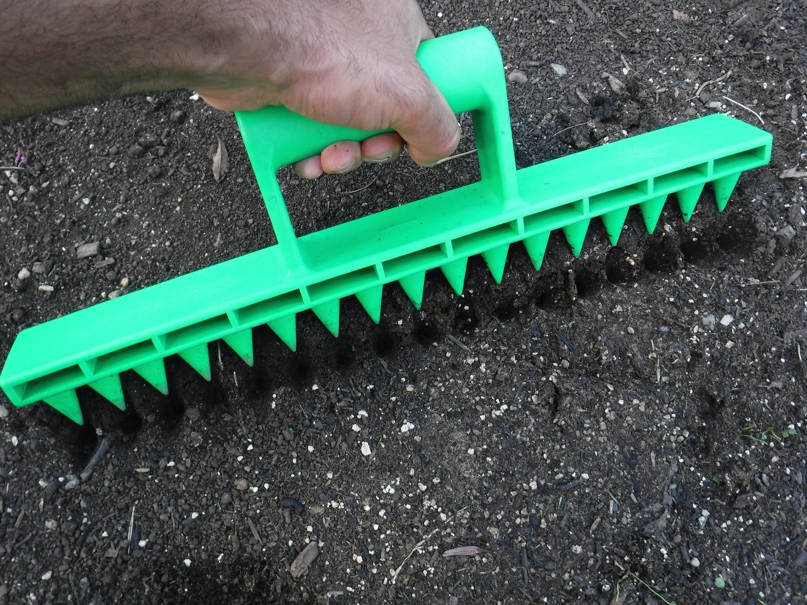 Hand using the green plastic rake tool to dig holes in soil