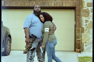A couple embracing in front of their garage, each is holding a gun which they display for the camera