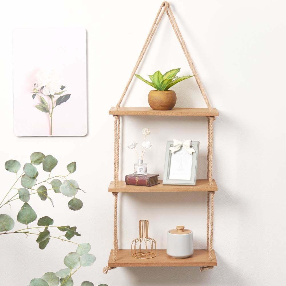 A three-tiered wooden wall shelf held together by thick rope. It houses a plant, photo frame, small books, and other decorative pieces.