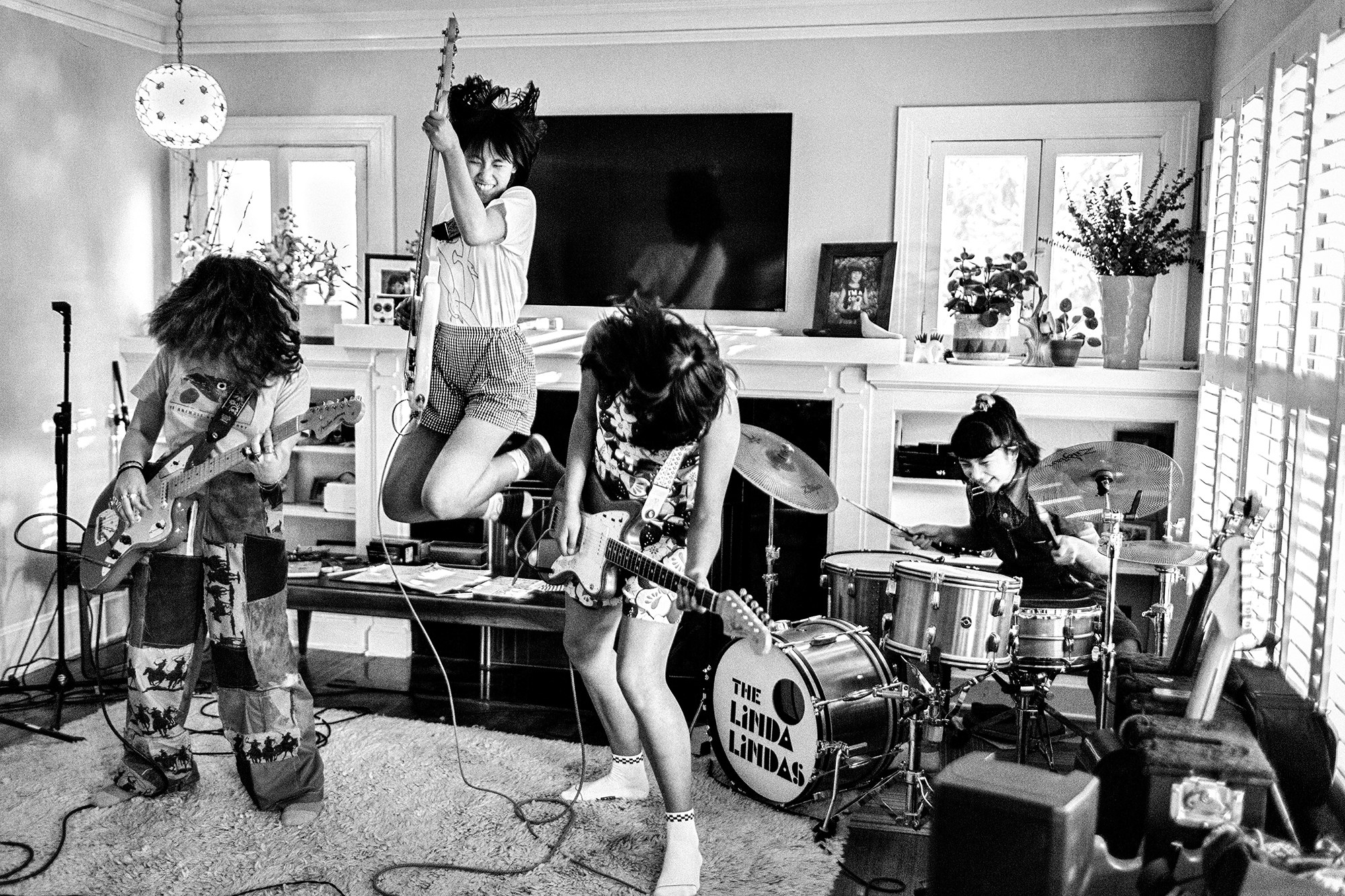 Four young girls jam out in a suburban living room, two playing guitar, one playing guitar and jumping into the air, and one on drums