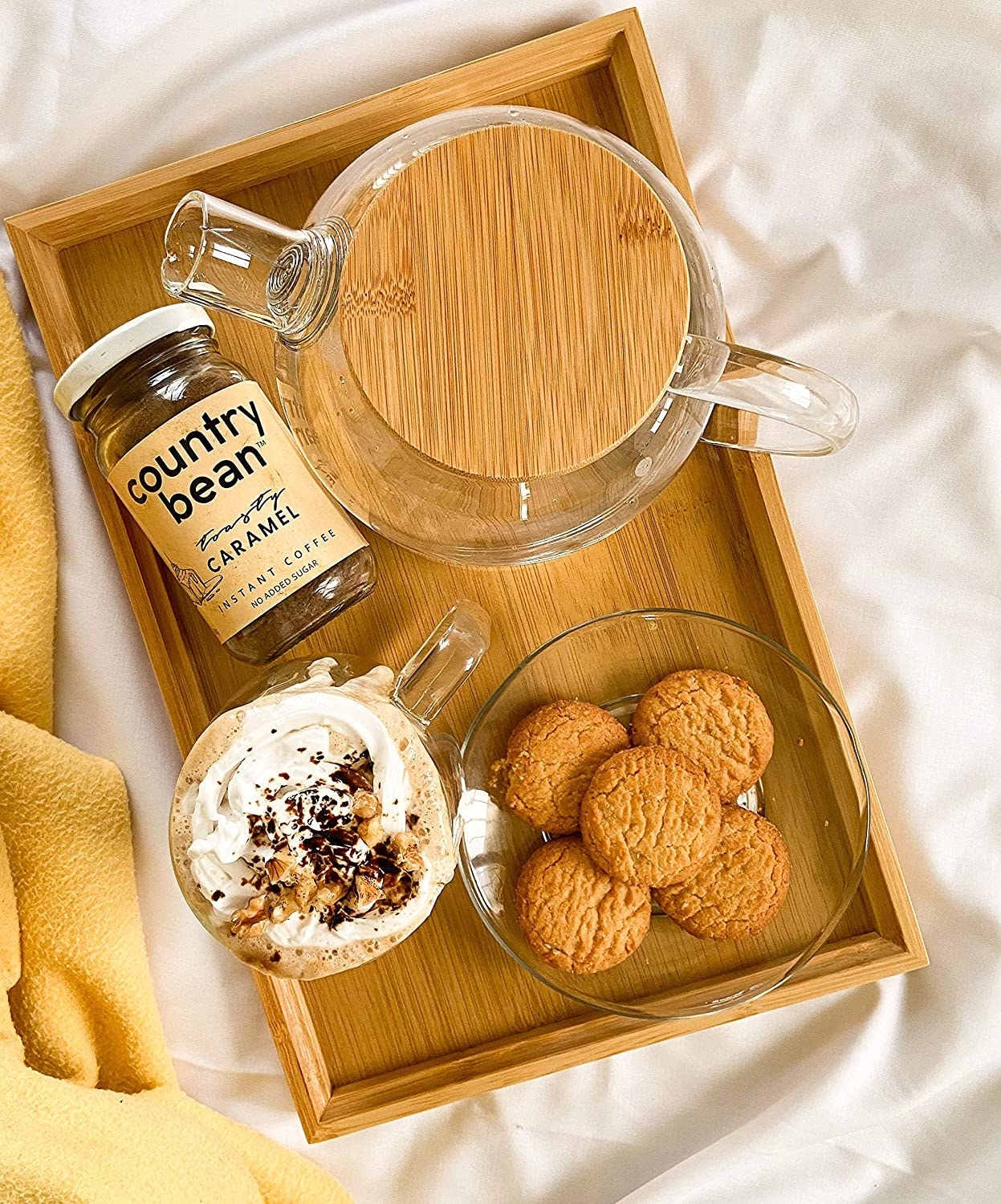 The coffee jar pictured with a cup of brewed coffee, a plate of biscuits, and a glass kettle. All of this is placed on a wooden tray.