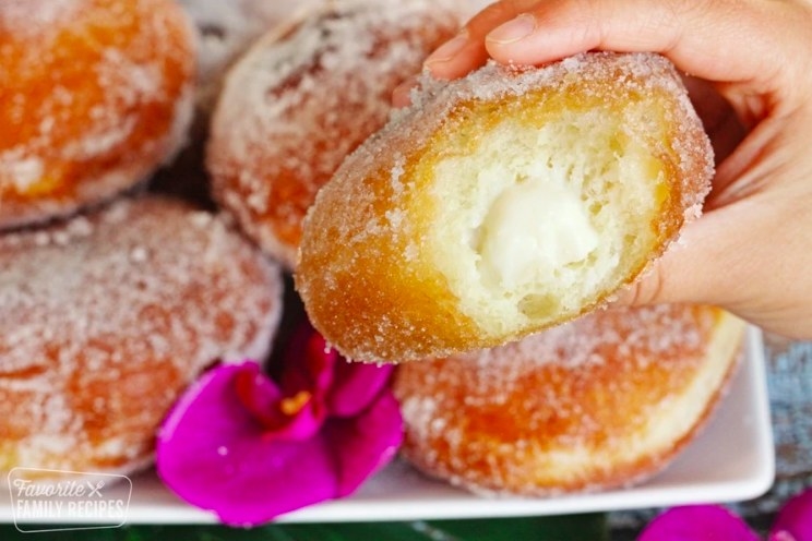 Hand holds up a Malasada donut with a bite taken out of it, revealing the filling inside
