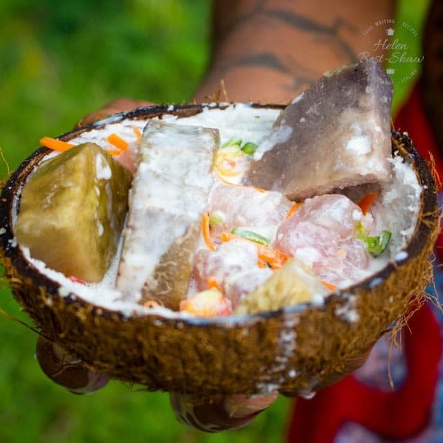 A hand holds out a halved coconut shell filled with Poisson Riu drenched in coconut milk. 