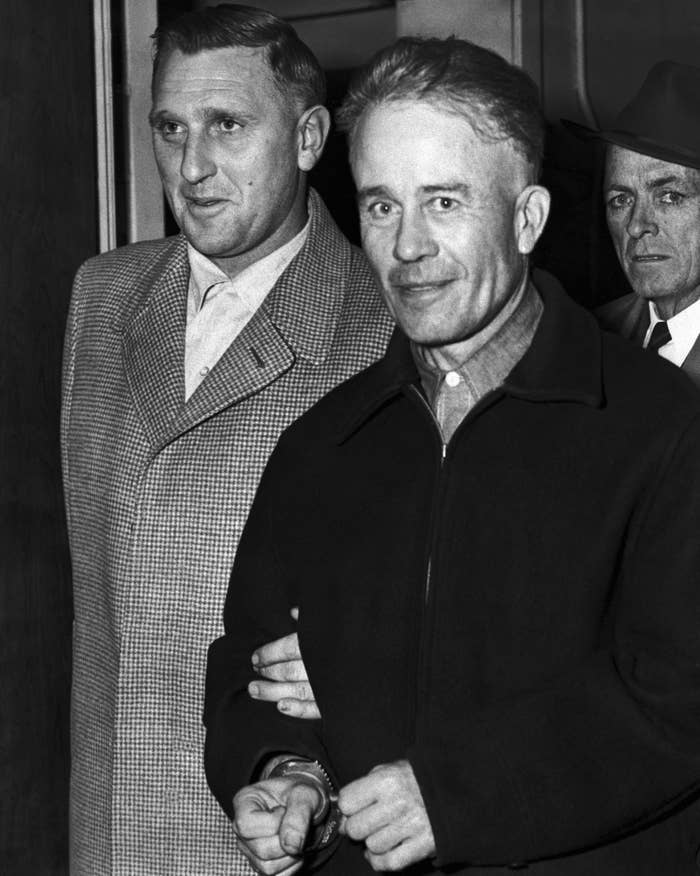 Ed Gein with a creepy smile on his face as he&#x27;s being escorted by police in handcuffs