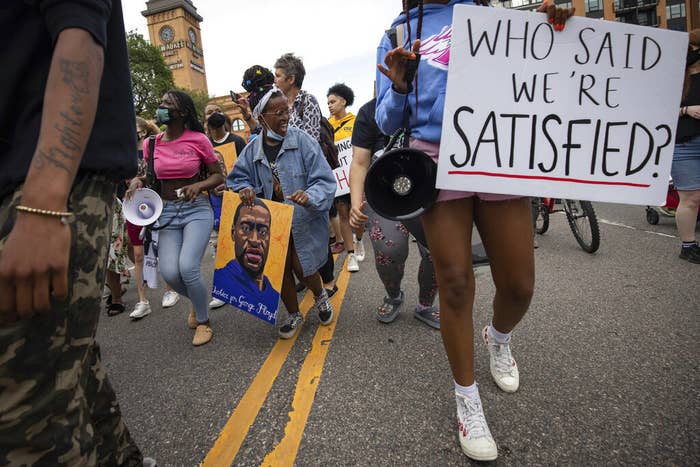 Protesters walking in the street carry megaphones, a painting of George Floyd&#x27;s face, and a sign reading &quot;Who said we&#x27;re satisfied?&quot;