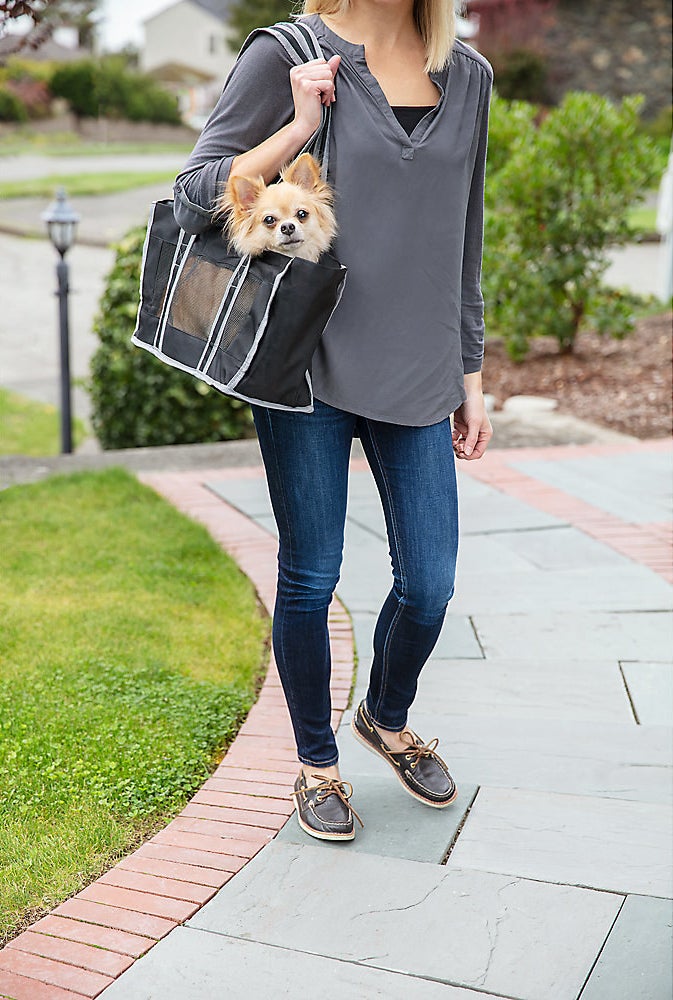 A small dog in a black bag with a person in blue, white and brown carrying it