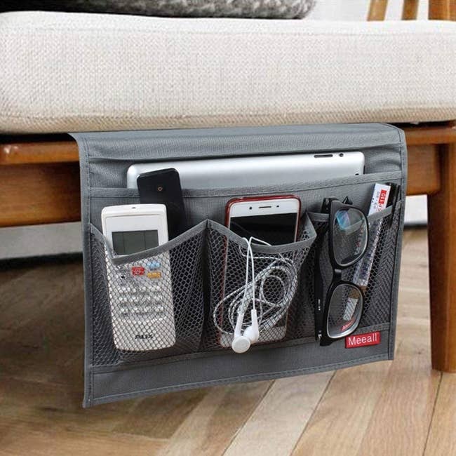 A gray fabric caddy tucked under a mattress with pockets holding a phone, remotes, glasses, and a laptop 