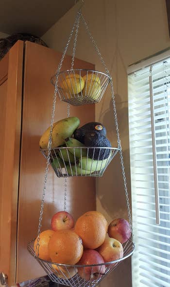 three tiered hanging metal baskets with fruits in them hanging from a kitchen wall 