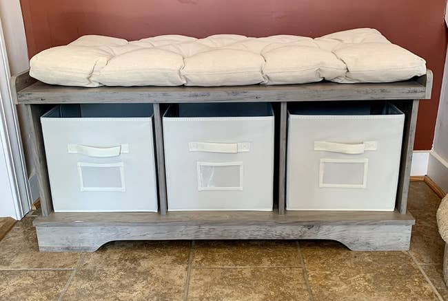 The bench with a cream-colored cushion and three matching storage bins underneath