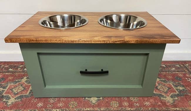 An elevated dog feeding station with a green drawer as a base and a flat wooden top with two silver food bowls installed 