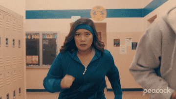 Asian woman wearing blue workout gear and a matching bandana walking really quickly trying to catch up with a person (off camera) down a high school hallway.