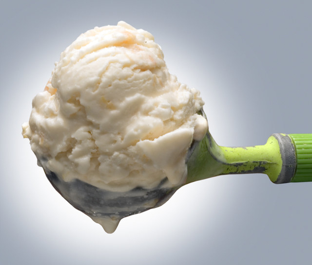 An ice cream scooper holds a single scoop of melting vanilla ice cream.