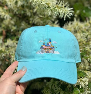 a teal hat with an embroidery of sleeping beauty castle on it in the clouds