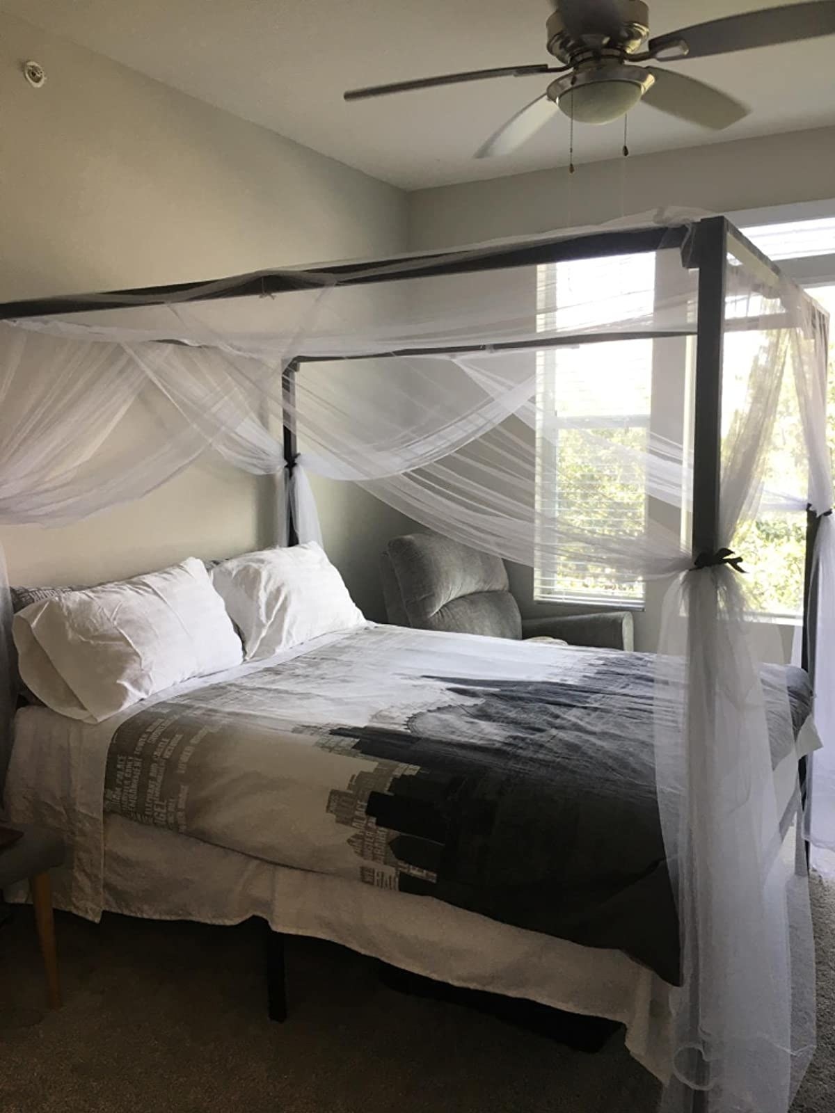 The canopy bedframe assembled in a bedroom. It has sheer curtains on it, tied to each of the four corners. It looks really nice in the small space!