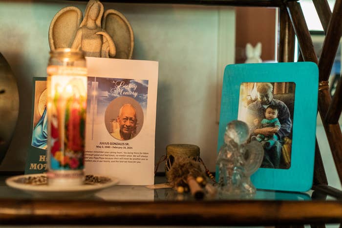 A shelf shows a small ceramic angel statue, a funeral program for Julius Gonzales Sr, a Lady of Guadalupe candle, and a picture of an older man holding an infant in his lap
