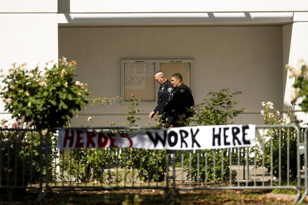 Two police officers walk behind a fence where a sign reading &quot;Heroes work here&quot; is posted