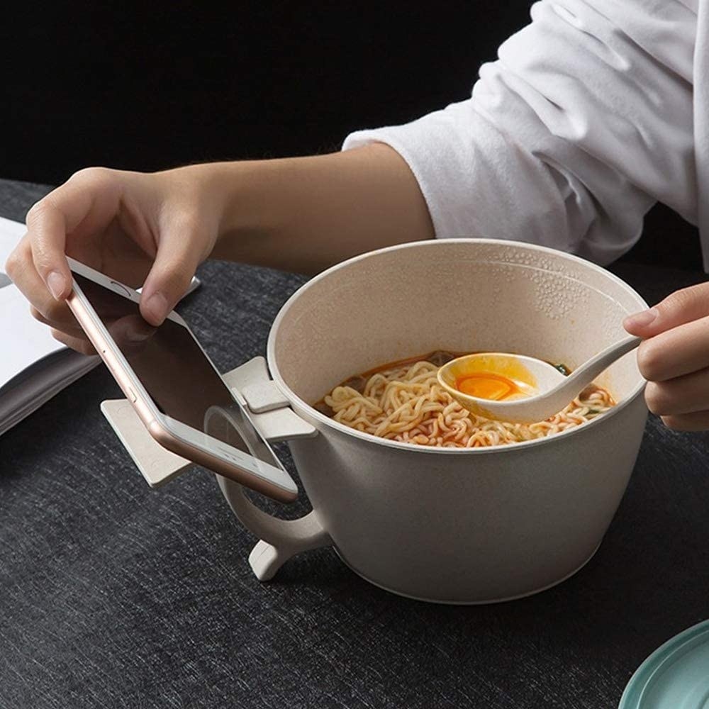 A person eat out of a large bowl  of ramen with a phone attached to the side
