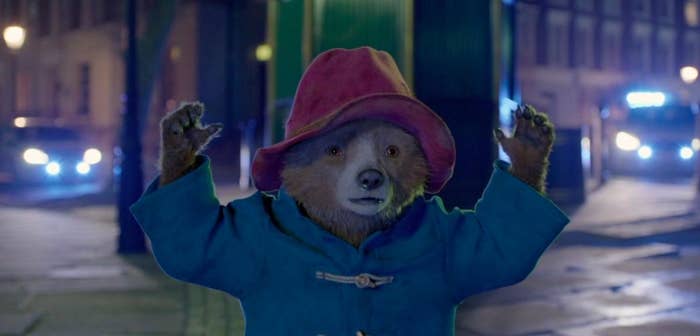 Paddington raises his hands in the air on a darkened street with cars behind him
