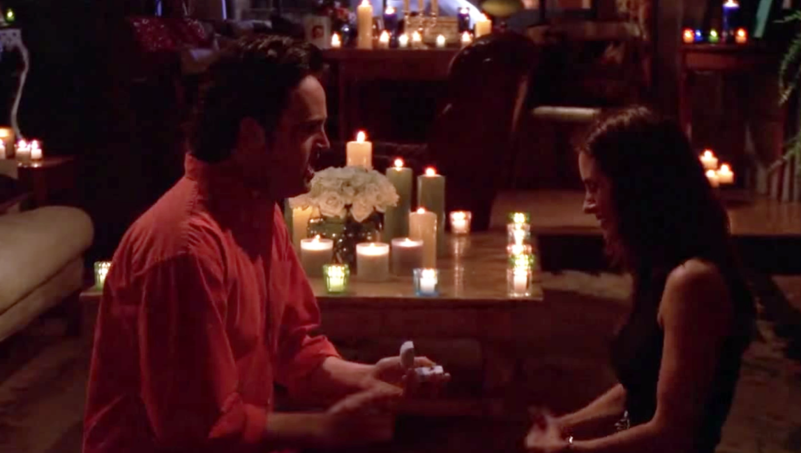 Chandler holds an open ring box to a shocked Monica while their kneeling around various candles.