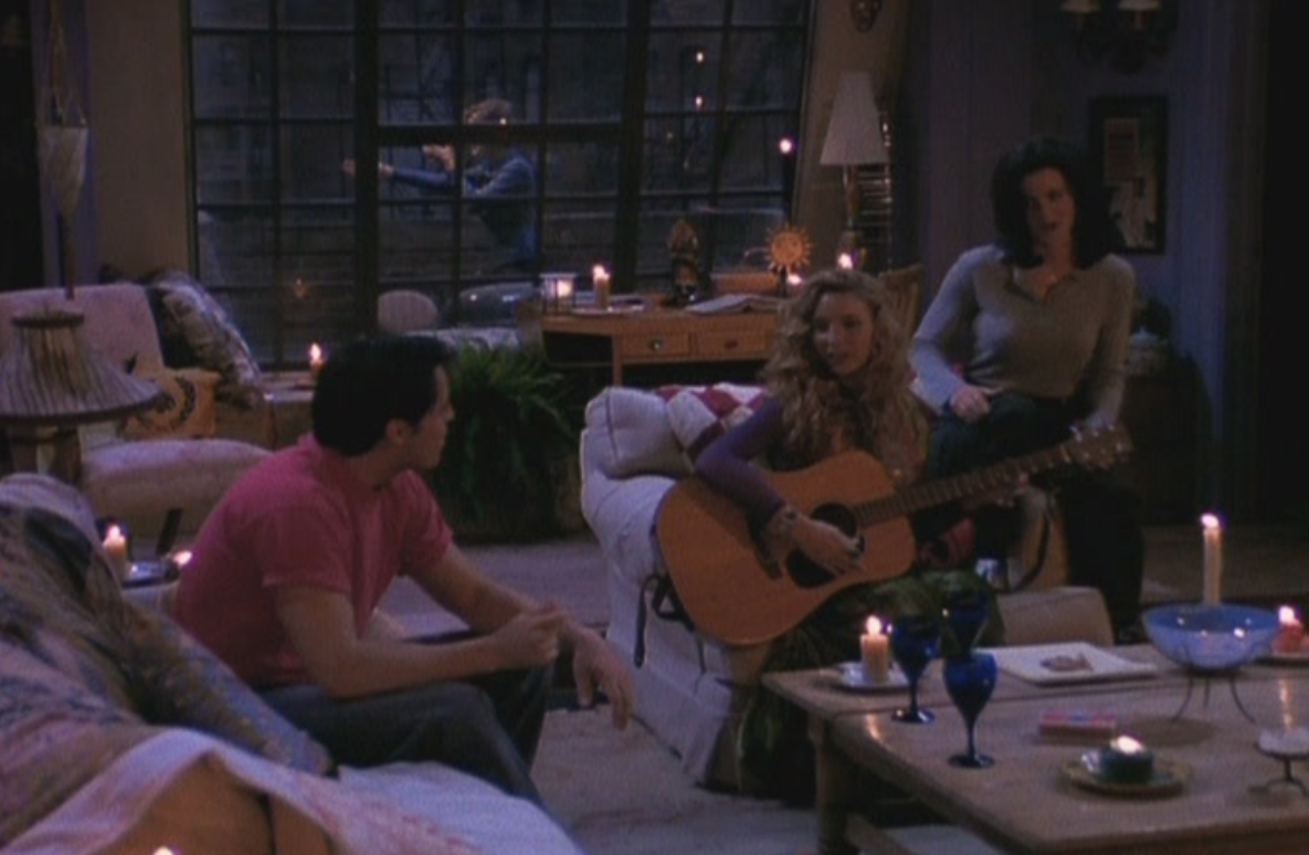 Monica, Phoebe, and Joey sit on Monic&#x27;s couches surrounded by various lit candles while Phoebe holds her guitar.