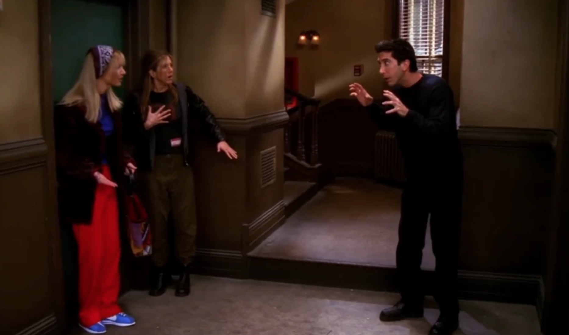Ross holds his hands out like claws to scare Rachel and Phoebe in the hallway of their apartment. 