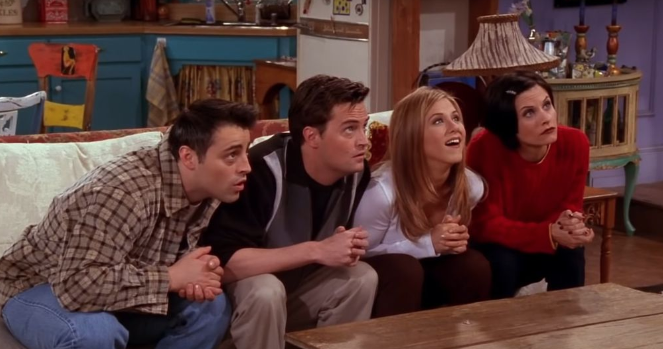 Joey, Chandler, Rachel, and Monica all sit on Monica&#x27;s couch with their hands folded and leaning forward expectantly.