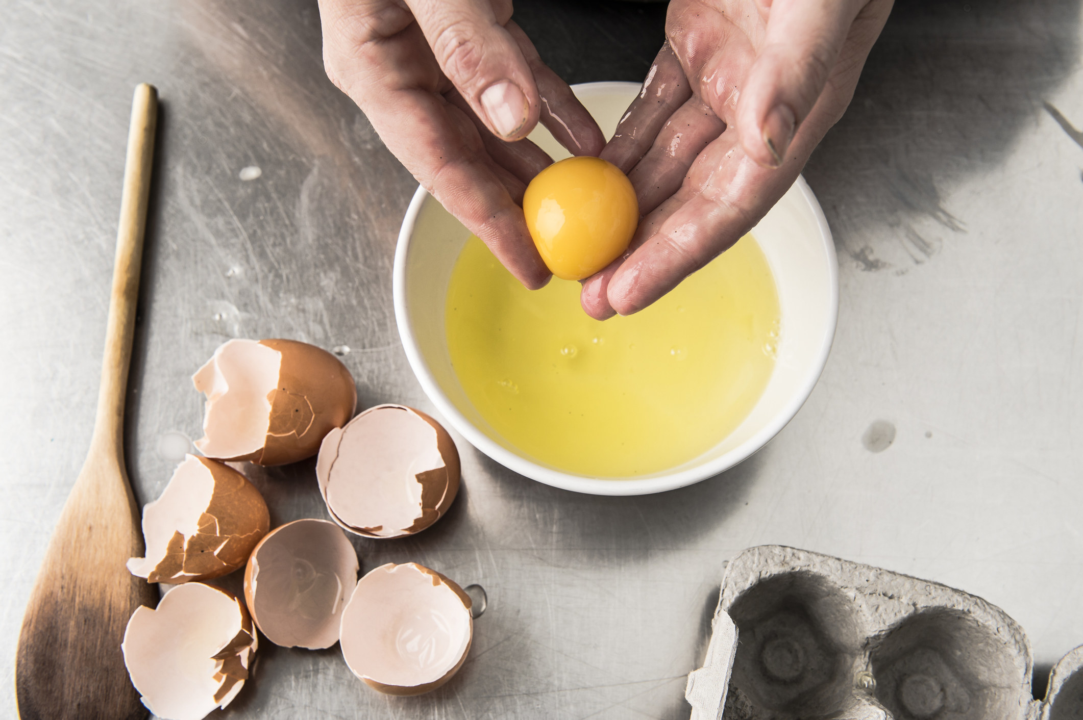 Hands holding an egg yolk over a bowl of beaten eggs.