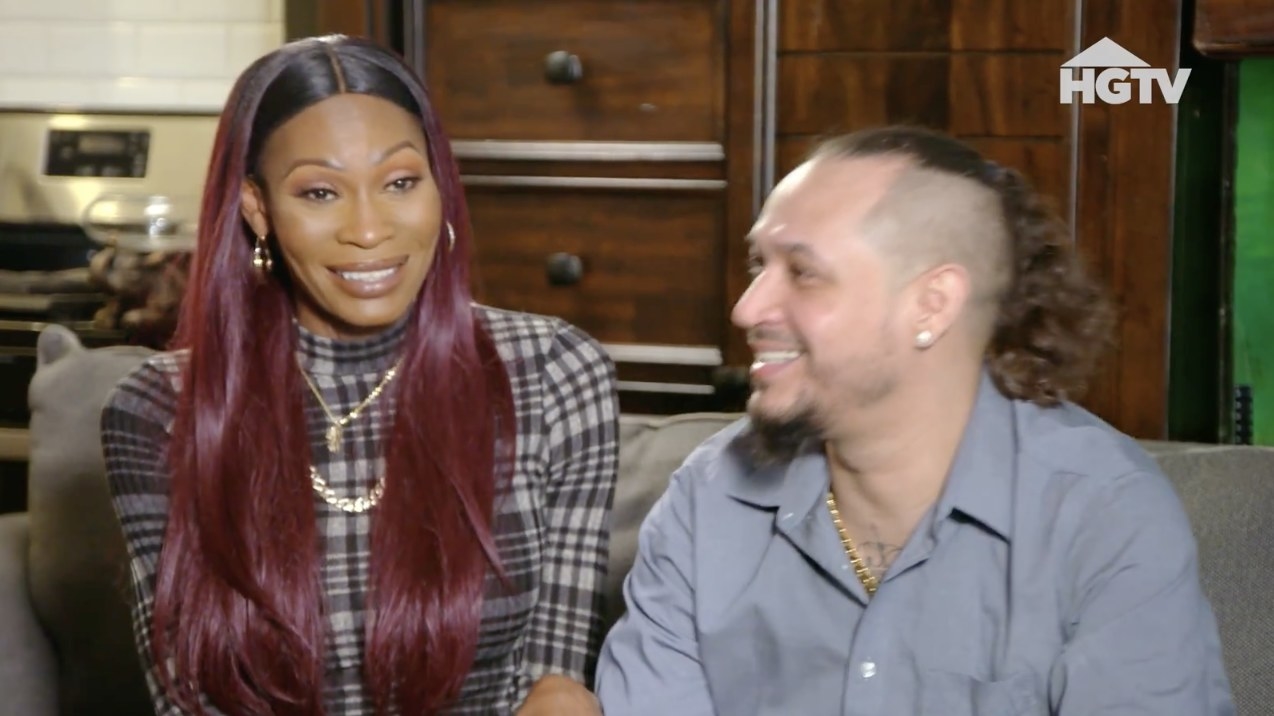 Dominique Jackson with long, straight hair sitting on a couch smiling with her fiancé, Edwin, a man with hair pulled back into a curly ponytail, shaved on the sides, and goatee, smiling