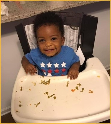 A young boy eats dinner in his high chair, making a mess with the food and smiling at the camera