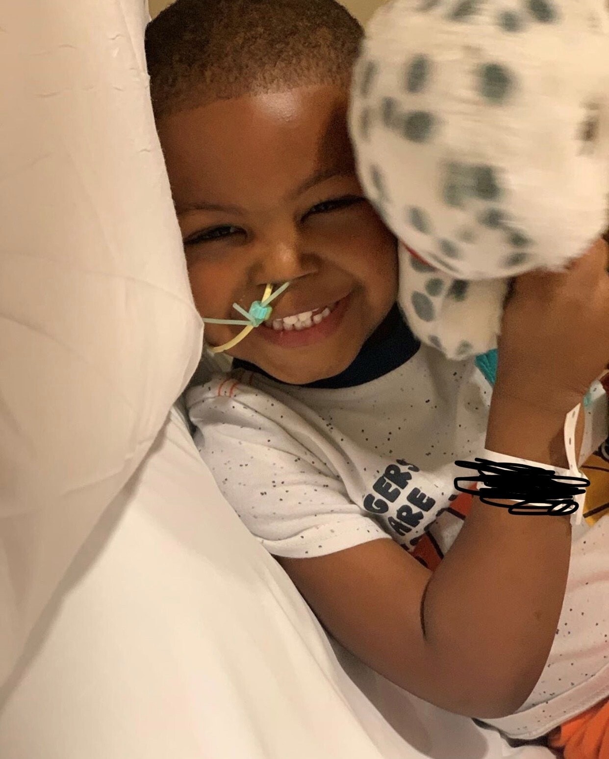 A young boy laughs while lying in a hospital bed, holding a stuffed animal 