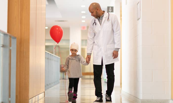 A child in a headscarf walks down a hospital hallway holding her doctor&#x27;s hand and a balloon
