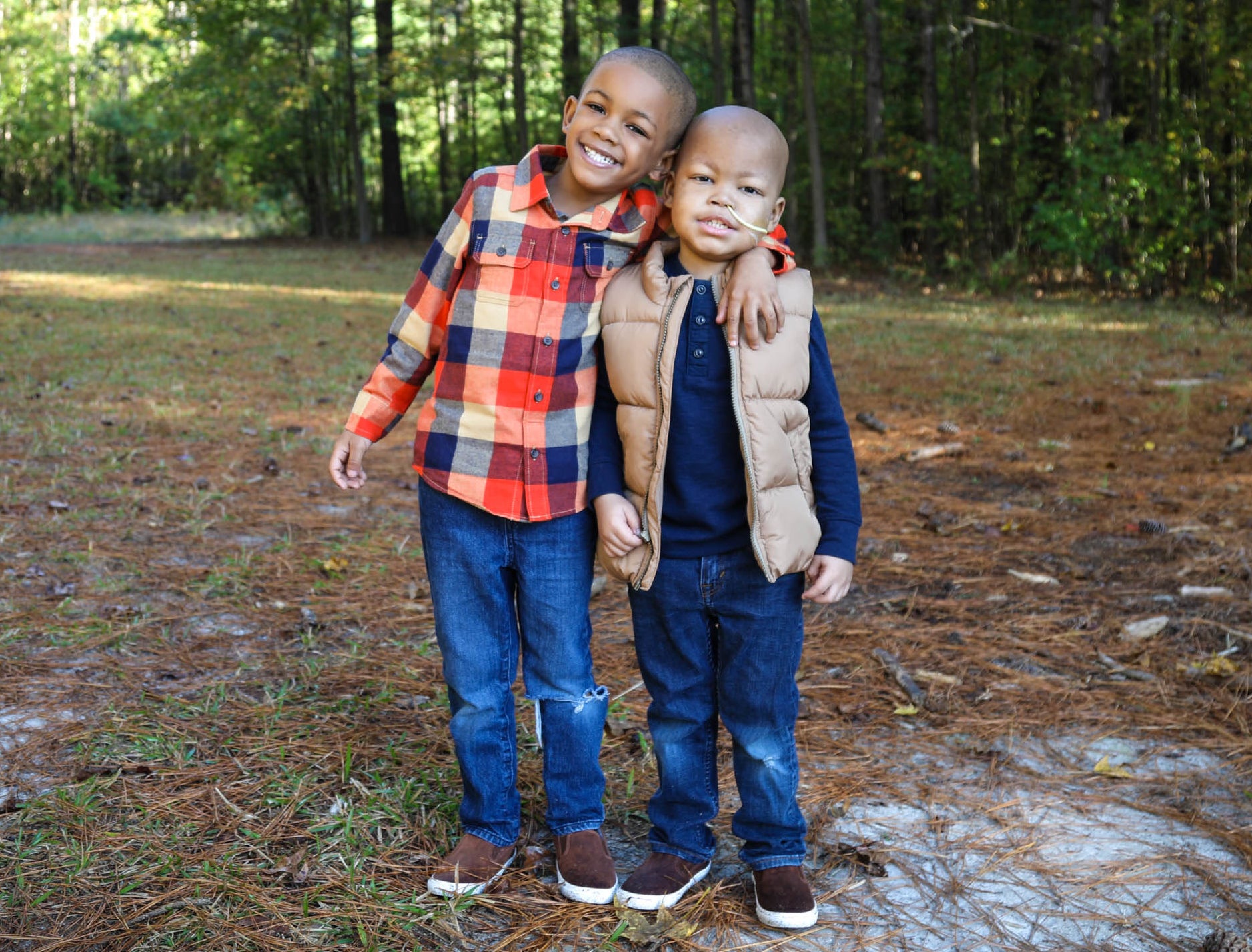 Two young boys smile with their arms around each other, standing in the woods
