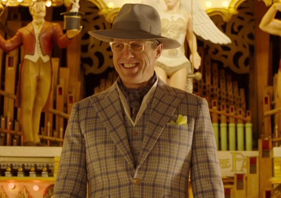 Hugh Grant as Phoneix; he smiles and wears a check suit, glasses and a hat, standing in front of an elaborate organ