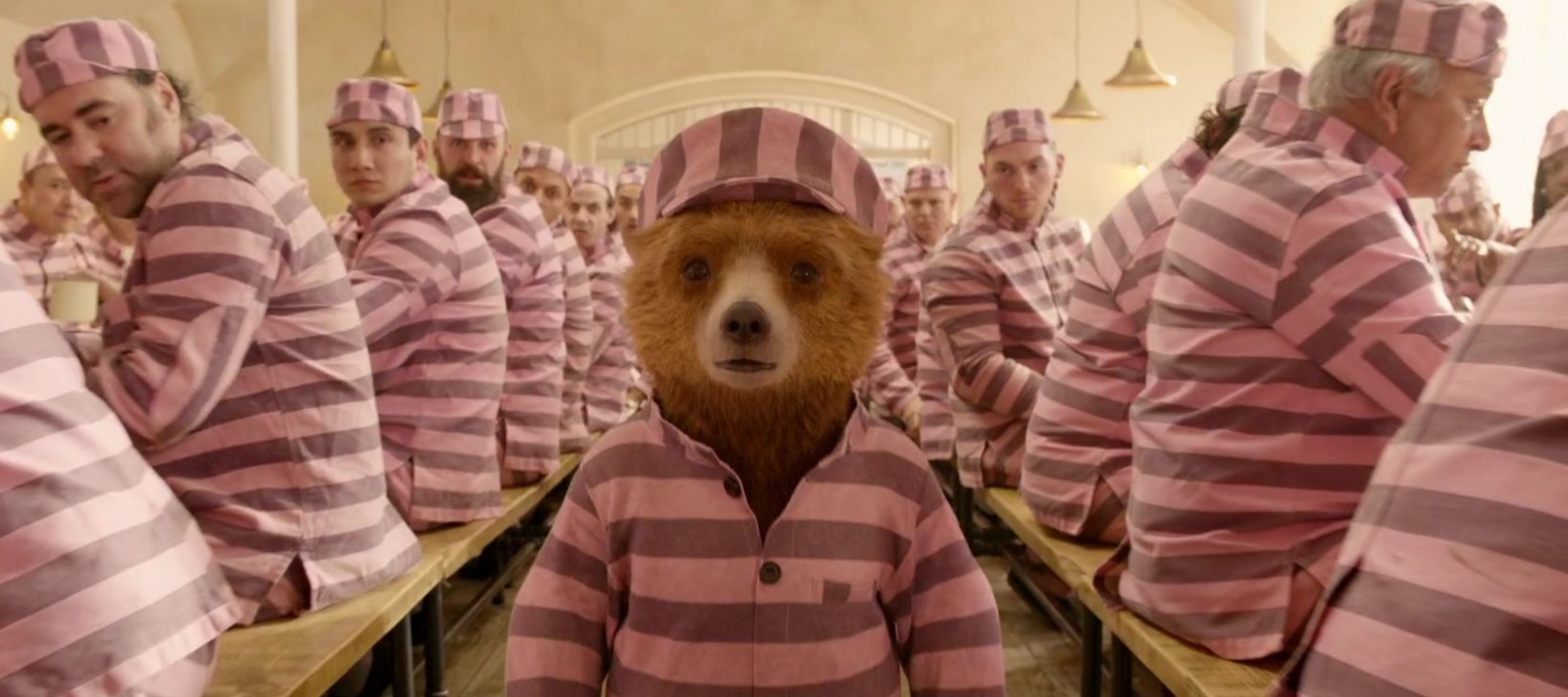Paddington stands between two rows of seated prisoners; they are all wearing pink striped prison uniforms