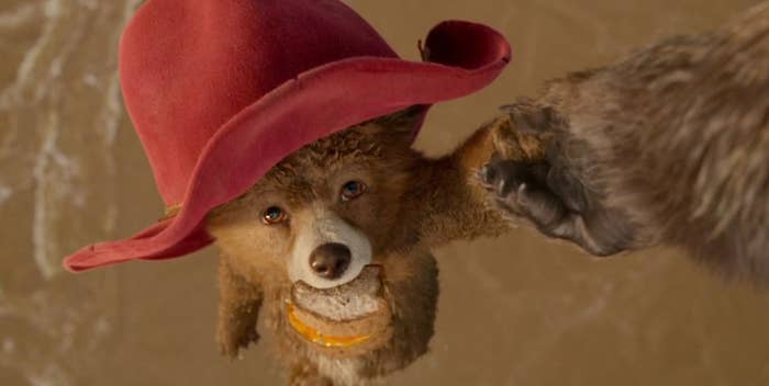 Young Paddington dangles over water wearing a red hat with a marmalade sandwich in his mouth