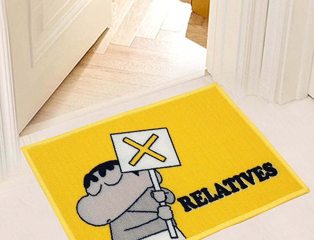 A bright yellow doormat with a monochrome illustration of Shinchan holding a board with a cross on it and the word &#x27;Relatives&#x27; written just underneath.