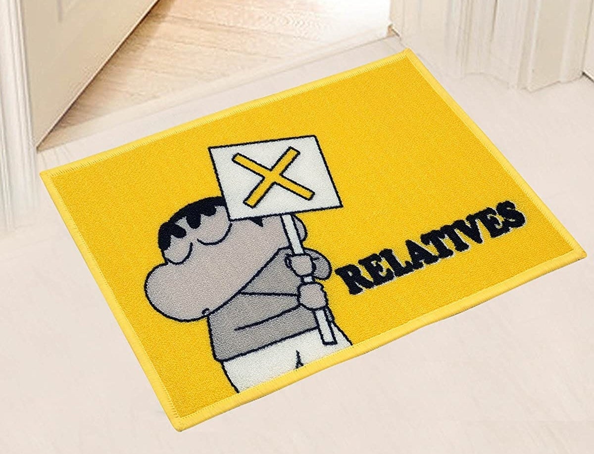 A bright yellow doormat with a monochrome illustration of Shinchan holding a board with a cross on it and the word &#x27;Relatives&#x27; written just underneath.