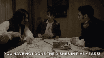 Three dark-haired vampires in ruffled shirts, one with a vest and two in coats, sitting around a table, the one in the vest says You have not done the dishes in five years and slams the table with his index finger