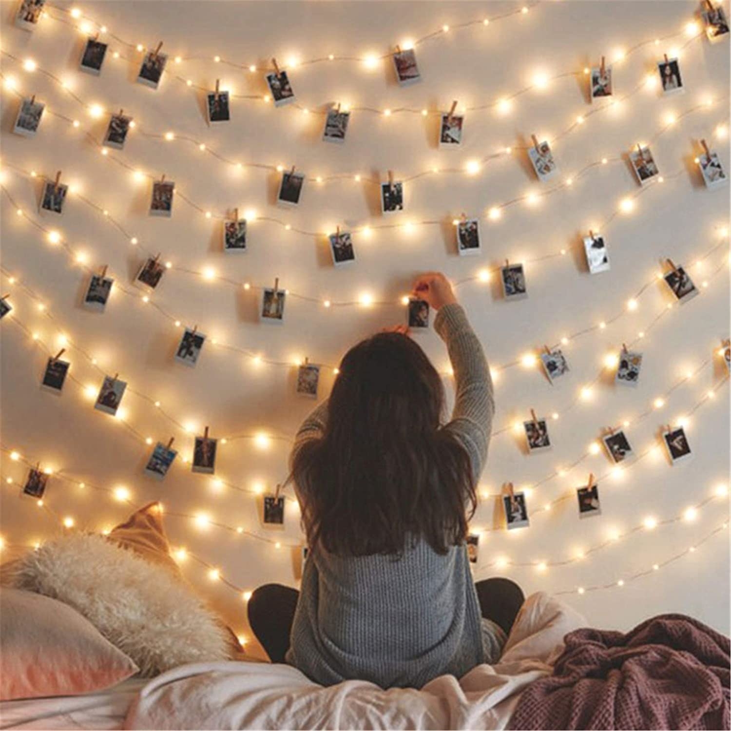 A wall of fairy lights with instant photos clipped onto the fairy lights with small wooden pegs. A woman&#x27;s back can be seen, she is clipping another photo onto the lights.
