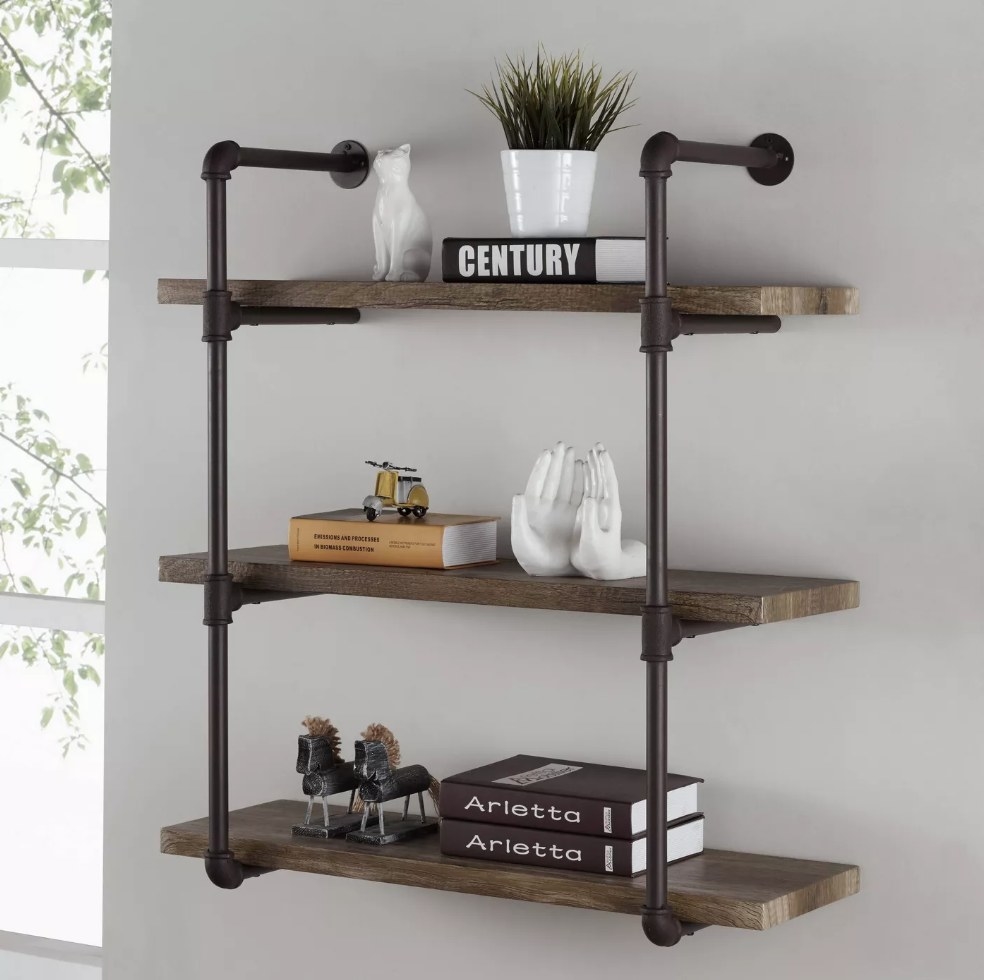 A three-tier industrial pipe shelf displayed on a wall and filled with books, a plant, and other decorative items